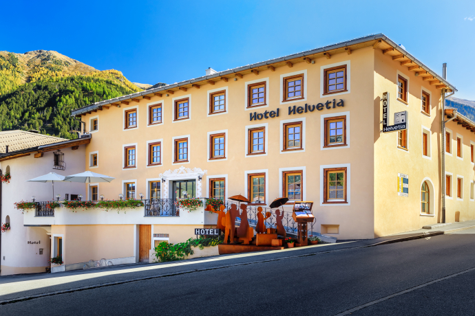 Hotel Helvetia in Müstair mit Terrasse und Sitzplätzen vor alpiner Kulisse.